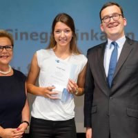 Drei Personen bei einer Preisverleihung; die Frau in der Mitte hält ein Zertifikat in der Hand; sie lächeln alle.