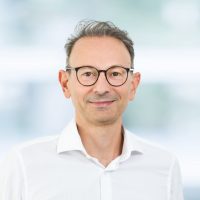 Mann mit Brille und weißem Hemd lächelt vor unscharfem Hintergrund.