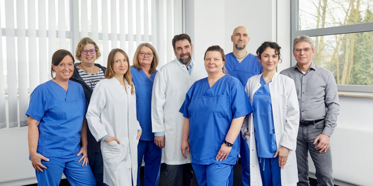 Eine Gruppe aus acht Medizinern und Pflegern in blauer und weißer Kleidung steht in einem hellen Raum zusammen.