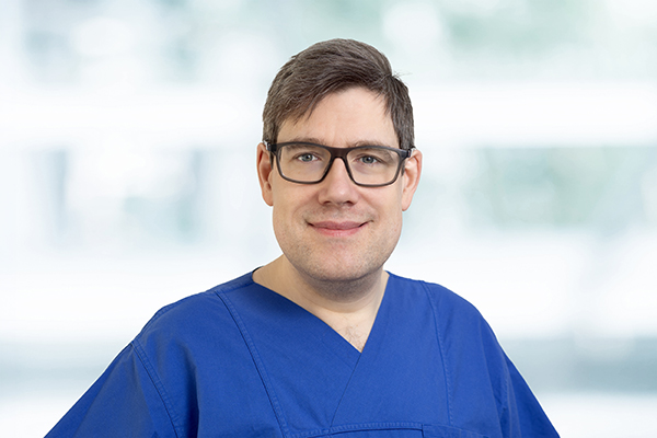 Mann mit Brille in blauer medizinischer Kleidung lächelt vor unscharfem Hintergrund.