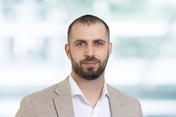 Ein Mann mit Bart in einem beigen Sakko und weißem Hemd vor unscharfem Hintergrund.