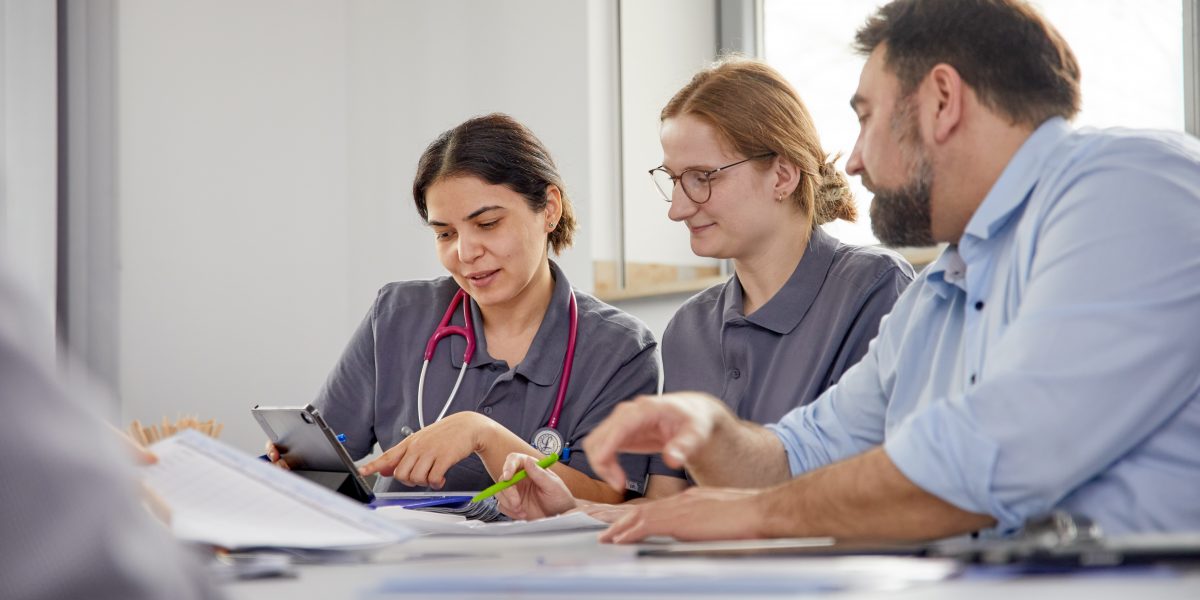 Drei Personen in medizinischer Kleidung, diskutieren an einem Tisch. Eine Frau zeigt auf ein Tablet mit Stethoskop um den Hals.