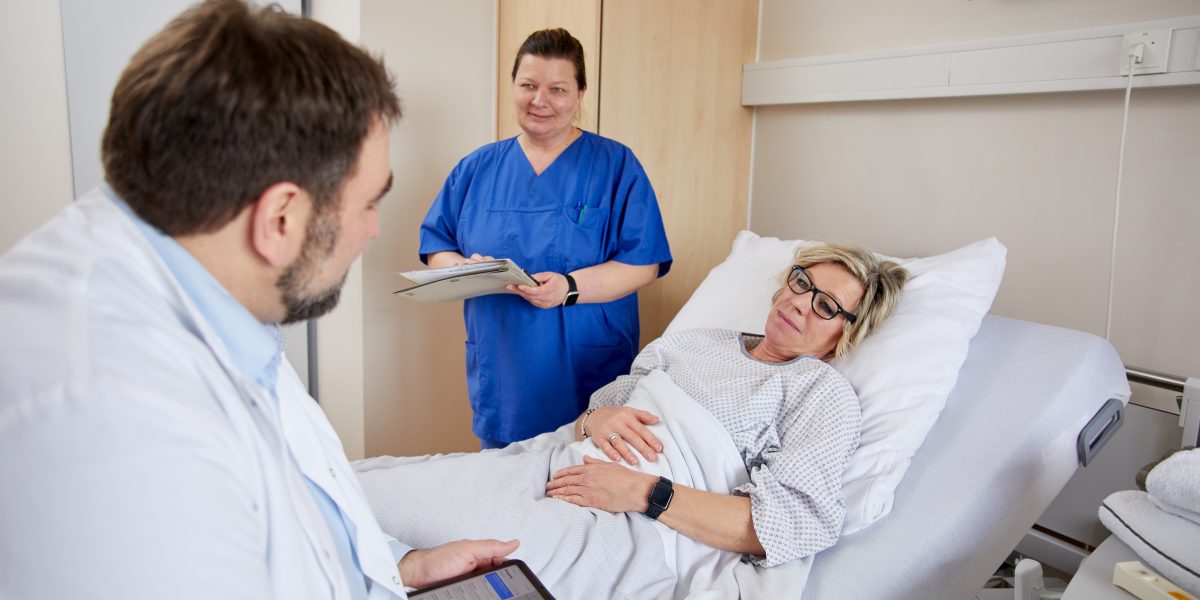 Arzt und Krankenschwester sprechen mit einer Patientin im Krankenhausbett. Die Krankenschwester hält einen Notizblock.