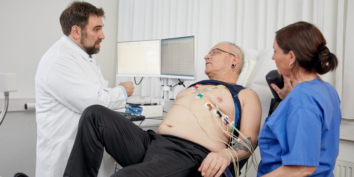 Doktor spricht mit einem Patienten beim EKG, während eine Krankenschwester assistiert. Patientenbrust mit Elektroden verkabelt.