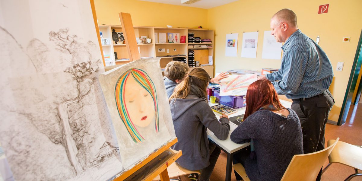 Lehrer unterrichtet eine kleine Gruppe Jugendlicher in einem Kunstzimmer mit Zeichnungen und Malutensilien.