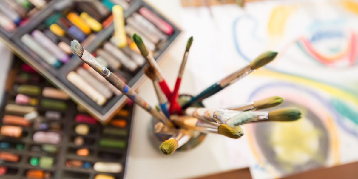 Malutensilien: Pinsel im Glas, bunte Pastellkreiden und Zeichnung auf einem Tisch.