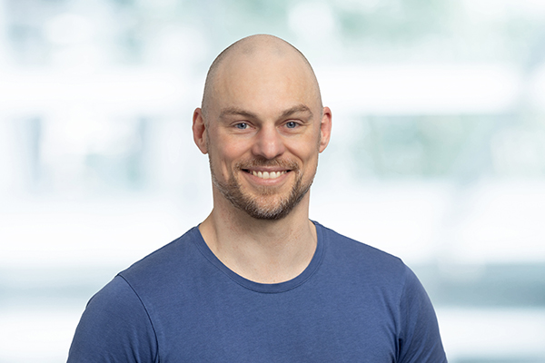 Ein lächelnder Mann mit Glatze und Bart, trägt ein blaues T-Shirt vor einem unscharfen, hellen Hintergrund.