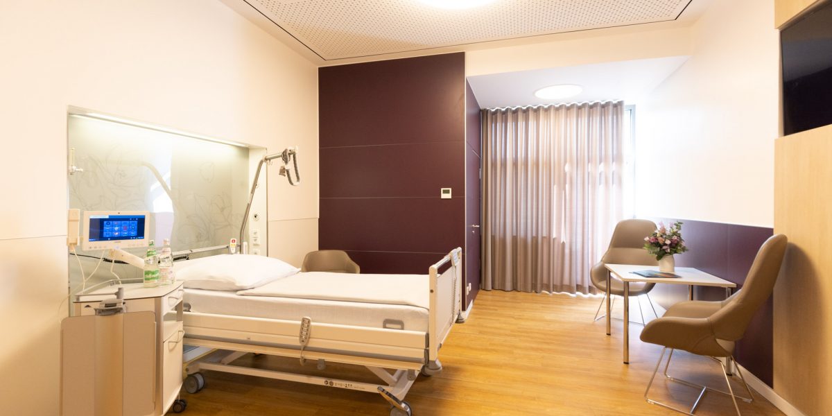 Helles Krankenhauszimmer mit Bett, medizinischer Ausrüstung, zwei Stühlen an einem Tisch und Blumen auf Holzfußboden.