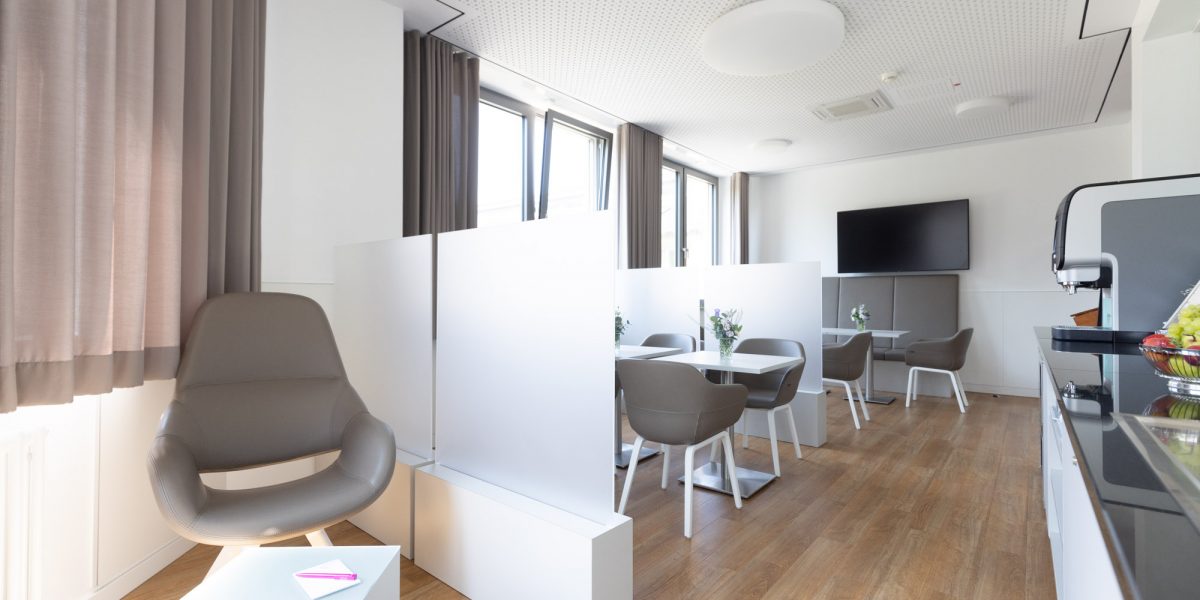 Moderner, heller Aufenthaltsraum mit grauen Stühlen, Tischtrennwänden und einem Fernseher an der Wand.