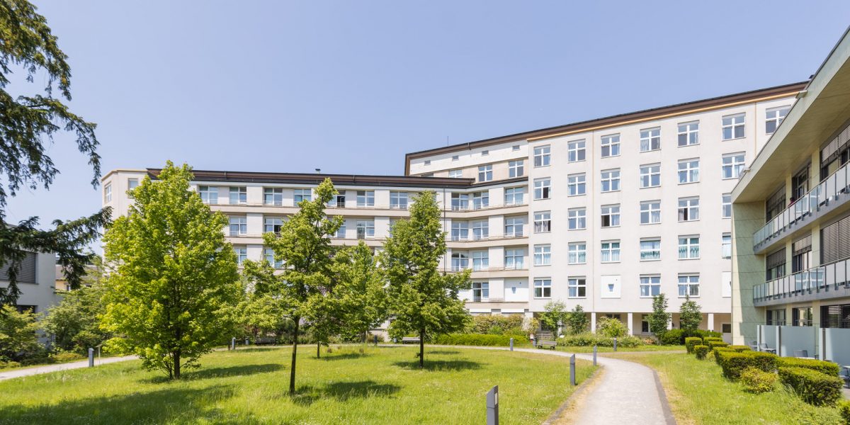 Mehrstöckiges Gebäude mit weißen Fassaden und blauen Fenstern vor grünem Rasen und Bäumen unter klarem Himmel.