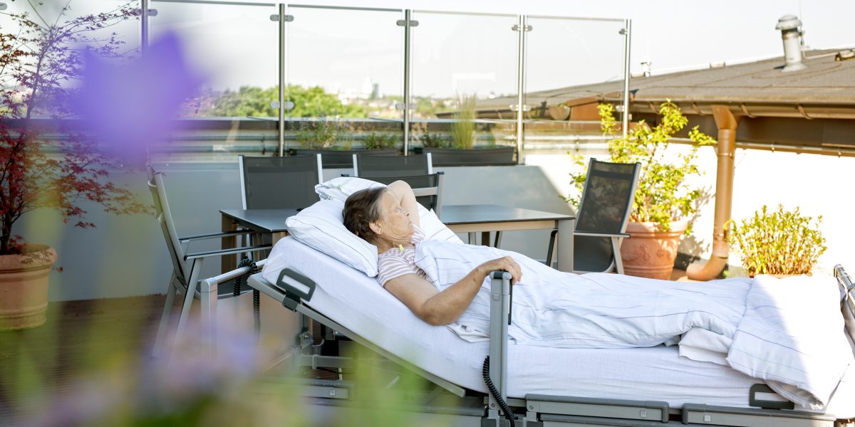 Ältere Person liegt in einem Krankenhausbett auf einer Terrasse, umgeben von Pflanzen und Gartenmöbeln.