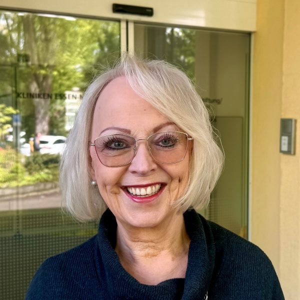 Person mit blonden Haaren und Brille lächelt vor einem Eingang mit Glasfront.