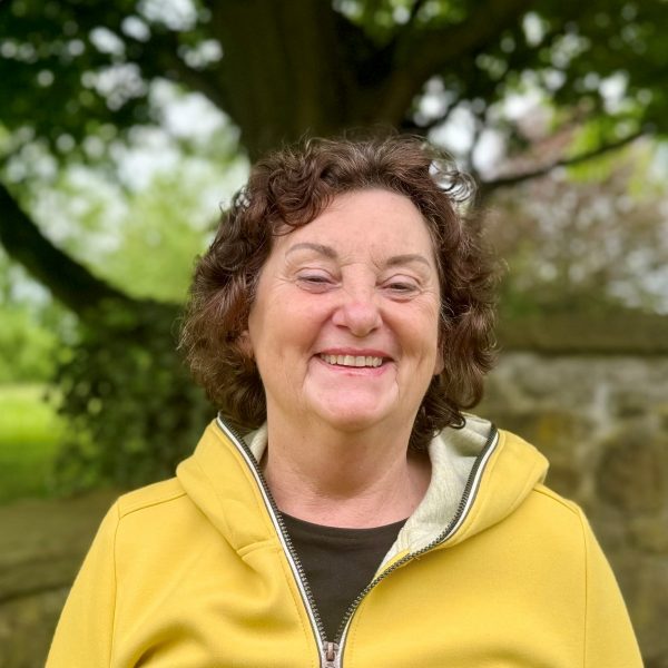 Frau mit lockigem Haar und gelber Jacke lächelt im Freien vor einem Baum und einer Steinmauer.