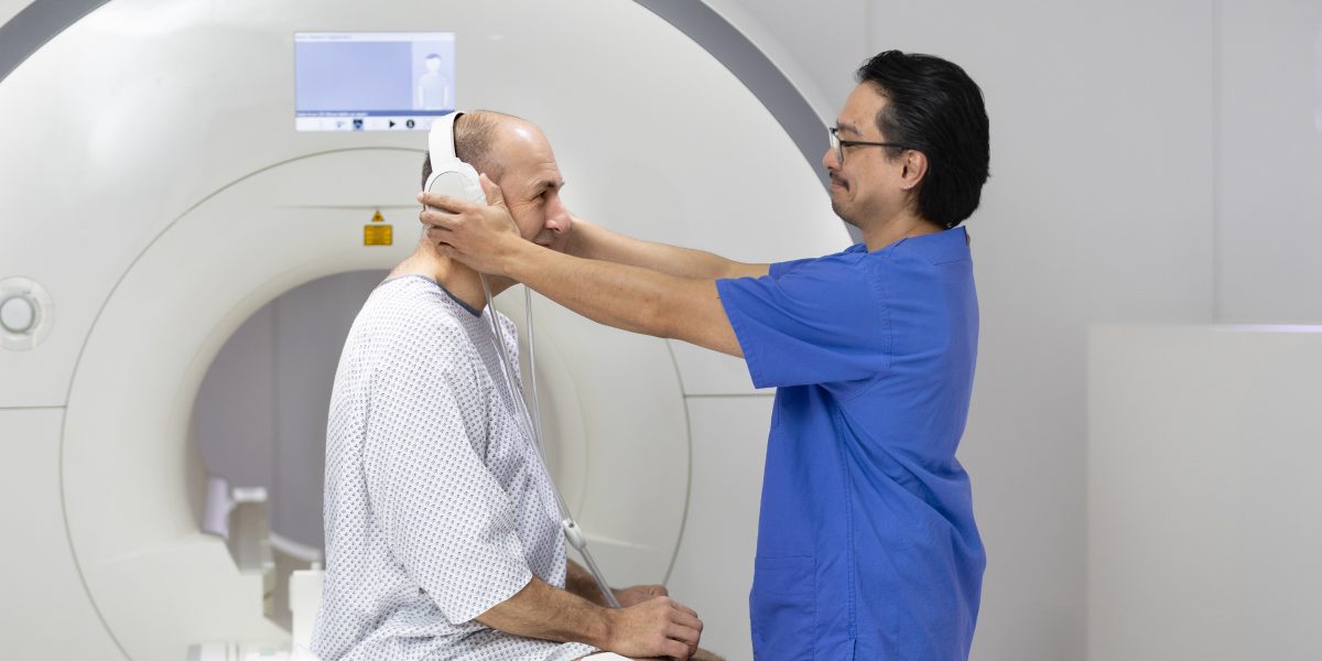 Ein Mann in Krankenhauskleidung bekommt Kopfhörer von einem Techniker vor einer MRT-Röhre angelegt.