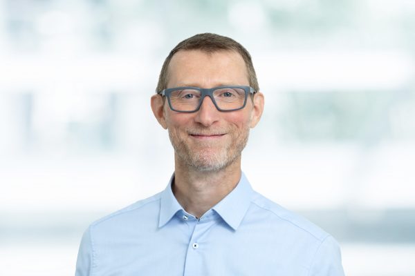 Porträt eines Mannes mit Brille und Bart in einem weißen Hemd, vor unscharfem Hintergrund.