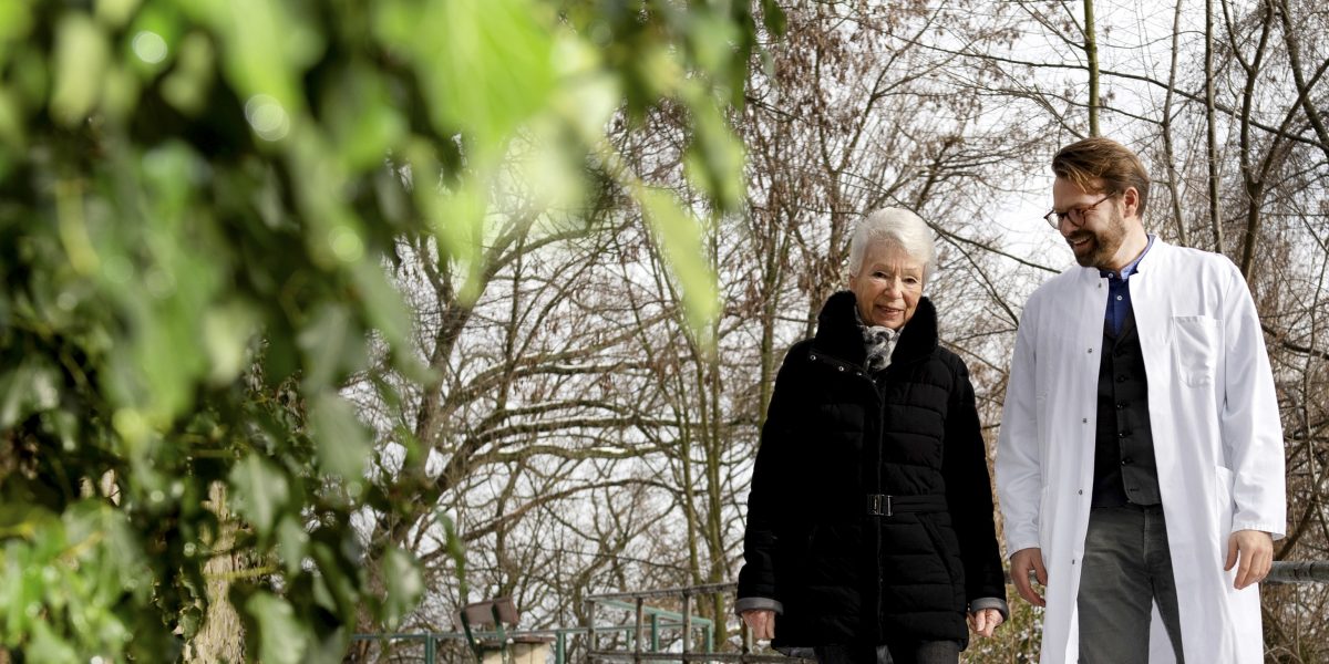 Ältere Frau und Arzt spazieren lächelnd im Freien. Links grüne Blätter, im Hintergrund winterliche Bäume.