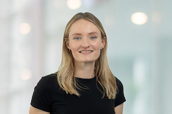 Junge Frau mit blonden Haaren lächelt und trägt ein schwarzes Shirt. Unscharfer Hintergrund mit hellen Lichtern.