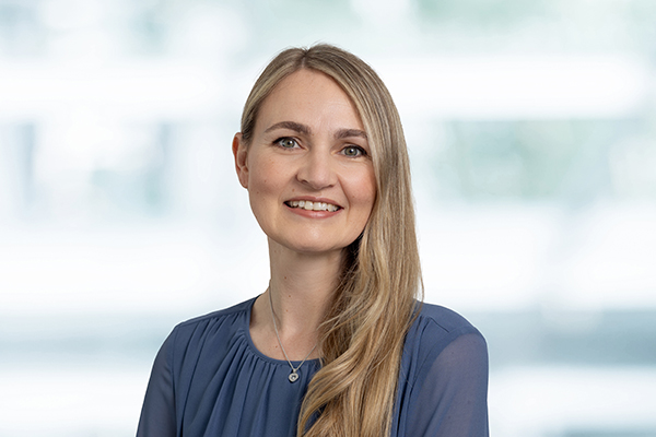 Eine lächelnde Frau mit langen blonden Haaren und blauer Bluse vor unscharfem Hintergrund.