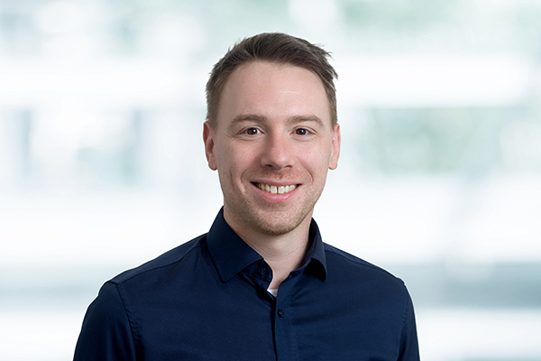 Porträt eines lächelnden Mannes in dunklem Hemd vor unscharfem, hellem Hintergrund.