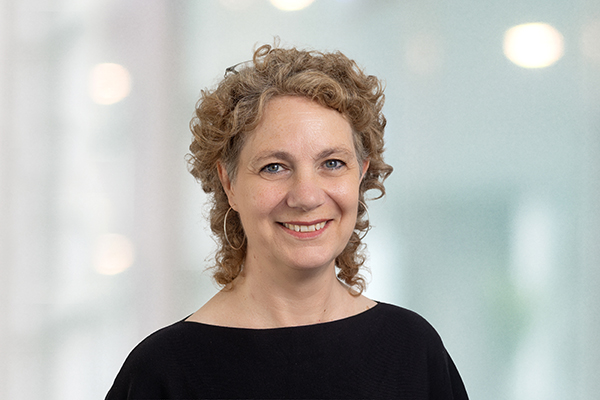 Eine Frau mit lockigem Haar lächelt in die Kamera vor einem unscharfen, hellen Hintergrund.
