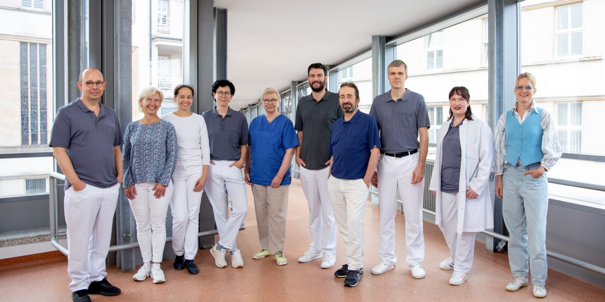 Gruppe von zehn Ärzten und Pflegern in einem Flur eines Krankenhauses, die in die Kamera lächeln.