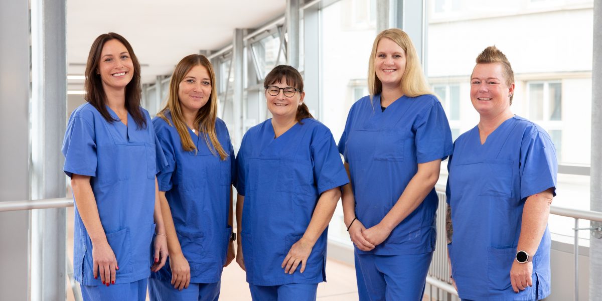 Fünf lächelnde Personen in blauen medizinischen Uniformen stehen in einem hellen Flur.