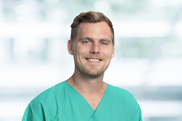 Ein lächelnder Mann in grüner medizinischer Kleidung vor einem unscharfen Hintergrund.
