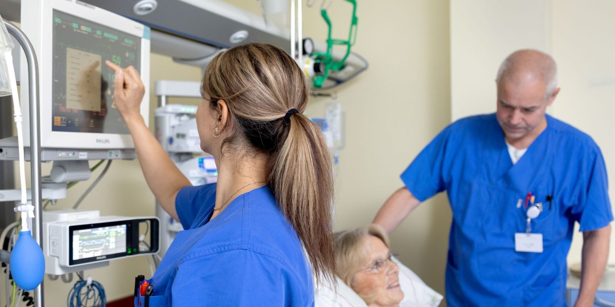 Two medical professionals in blue attire caring for an elderly patient in a hospital bed, monitoring screens.