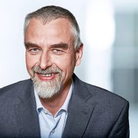 Mann mit grauem Bart und Anzug lächelt vor unscharfem Hintergrund.