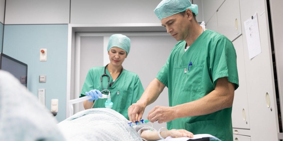 Two doctors in green surgical attire attending to a patient in the operating room. One holds a mask, the other performs a procedure. 