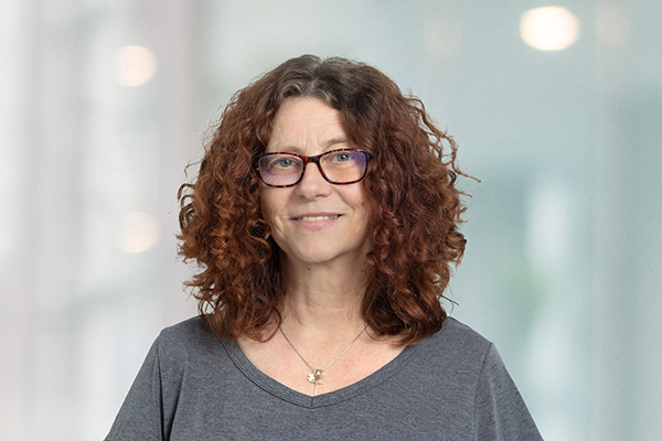 Frau mit lockigem, rotem Haar und Brille lächelt in die Kamera. Hintergrund ist verschwommen.