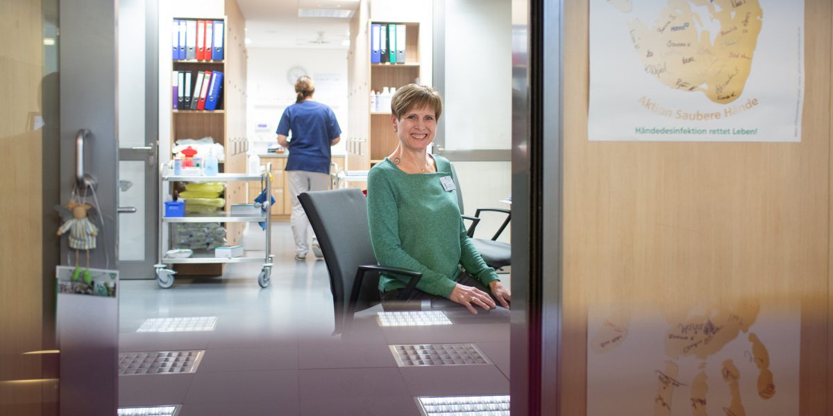 Frau am Schreibtisch, im Hintergrund Krankenpflegerin mit Pflegewagen. Plakat: 