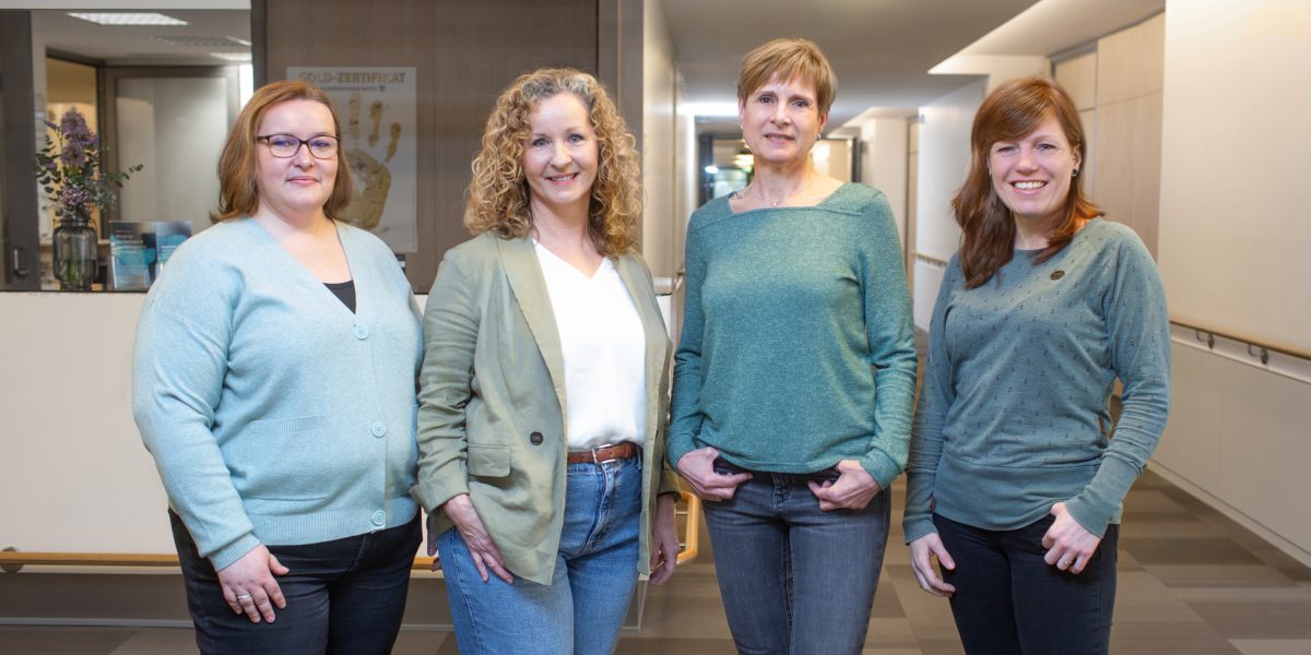 Vier Frauen stehen lächelnd in einem Flur, alle in grünen und blauen Tönen gekleidet.