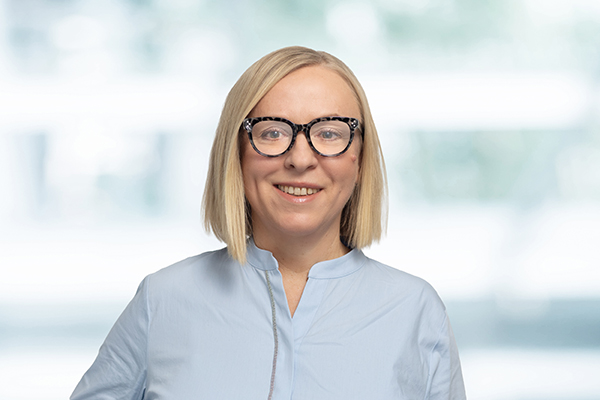 Frau mit blonder Bob-Frisur und Brille lächelt in die Kamera, trägt ein hellblaues Hemd. Verschwommener Hintergrund.