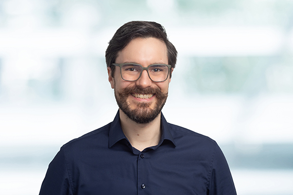 Ein lächelnder Mann mit Brille und Bart trägt ein dunkelblaues Hemd vor unscharfem Hintergrund.