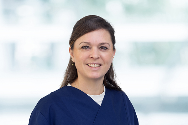 Frau mit dunkelbraunen Haaren lächelt in die Kamera. Sie trägt blaue Kleidung vor einem unscharfen, hellen Hintergrund.