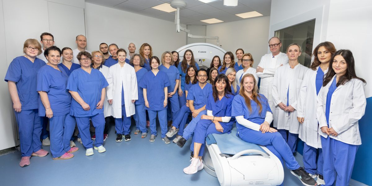 Große Gruppe von medizinischem Personal in blauer Kleidung und weißen Kitteln posiert in einem Behandlungsraum vor einem CT-Scanner.
