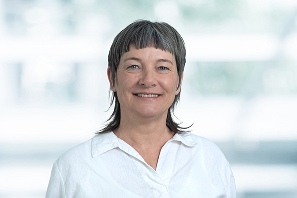 Porträt einer lächelnden Frau mit kurzen braunen Haaren und hellblauer Bluse vor neutralem Hintergrund.