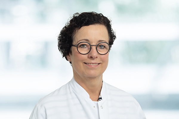 Person mit lockigem Haar und Brille, in weißem Hemd, lächelt vor unscharfem Hintergrund.