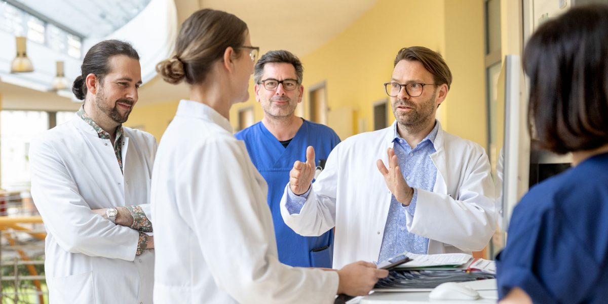 Ärzte in weißen Kitteln und blauen Scrubs diskutieren in einem Krankenhausflur. Ein Arzt gestikuliert erklärend.