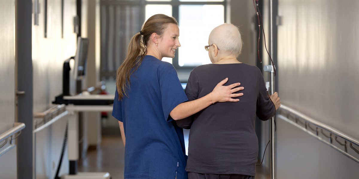 Pflegerin begleitet krebskranken Patienten mit Infusionsständer durch Krankenhausflur.