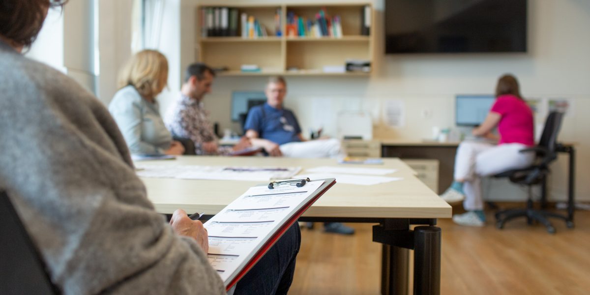 Vier Personen in einem Besprechungsraum, eine Person hält ein Klemmbrett, eine andere arbeitet am Computer.