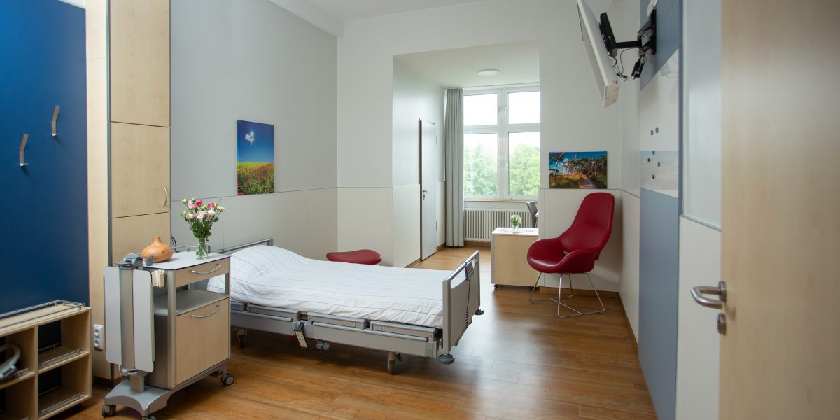 Helles Krankenhauszimmer mit Einzelbett, rotem Stuhl, Tisch und Fenster. Klare, moderne Einrichtung und Holzboden.