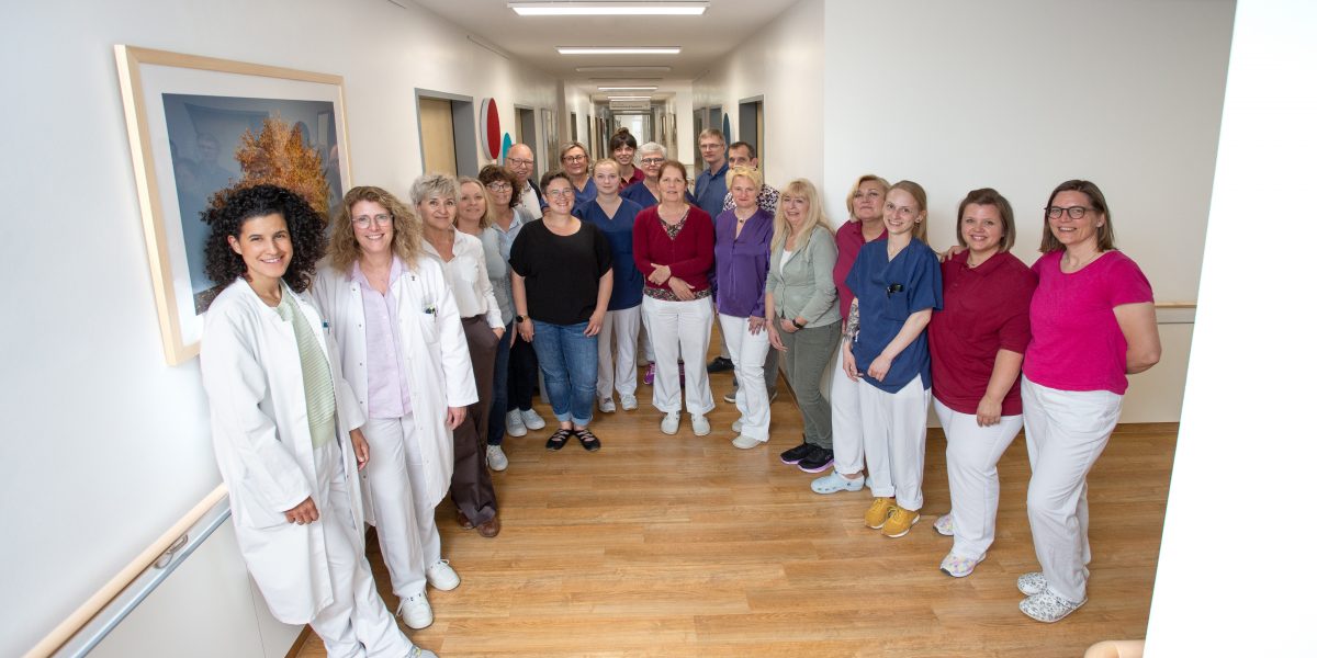 Gruppe von medizinischem Personal und Mitarbeitern in einem Krankenhausflur, lächelnd in die Kamera.