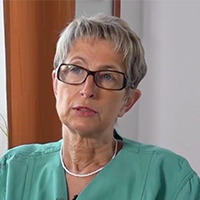 Ältere Frau mit grauem Haar und Brille in grünem Oberteil sitzt und schaut nachdenklich.