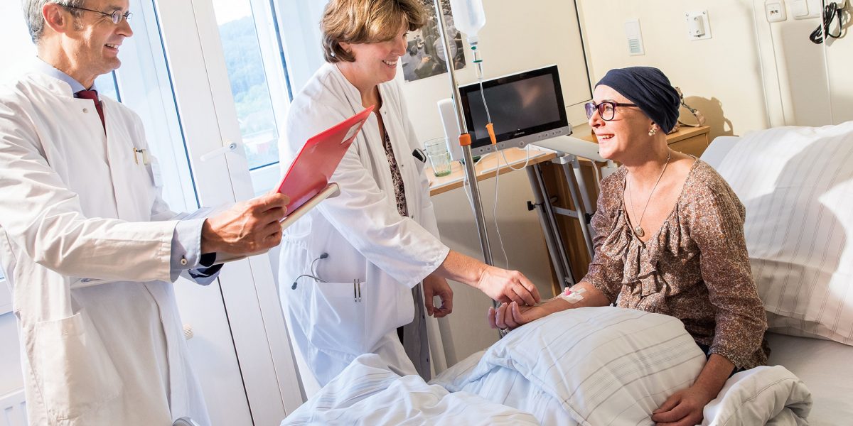 Ärzteteam kümmert sich lächelnd um patientin im Krankenhausbett. Sie wirkt positiv und unterstützt.