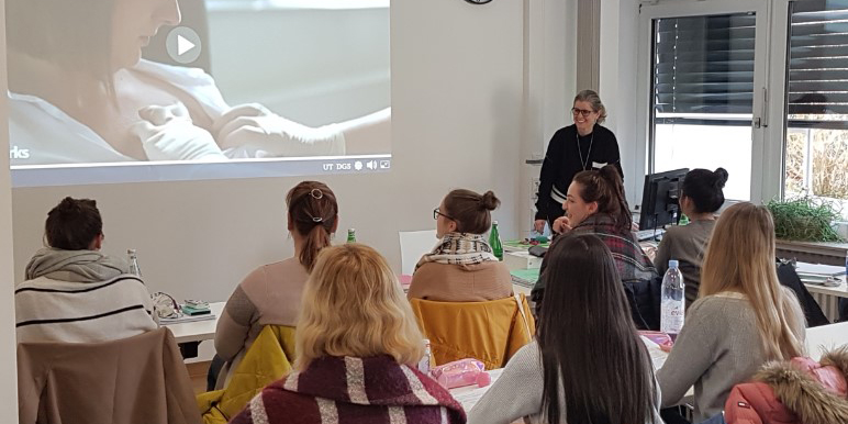 Eine Gruppe von Frauen schaut in einem Seminarraum ein Video auf einer Leinwand an, während eine Referentin spricht.