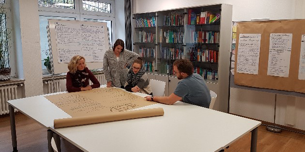 Vier Personen arbeiten in einem Konferenzraum an einem großen Papierbogen, umgeben von Präsentationstafeln und Bücherregalen.