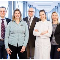 Fünf Personen in Geschäftskleidung stehen lächelnd in einem modernen Büroflur.