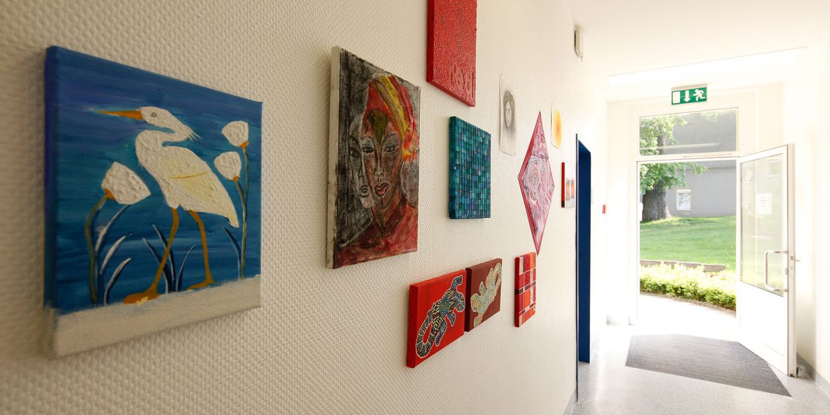 Gemälde hängen an einer Flurwand, darunter ein Vogelbild. Eine offene Tür führt zu einem sonnigen Garten.